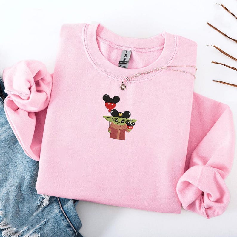 May include: A light pink sweatshirt featuring an embroidered design of a cartoon character holding a Mickey Mouse balloon. The character is wearing a red and brown outfit. A silver necklace with a sun pendant is on the sweatshirt.