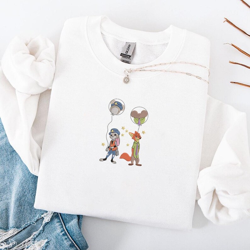 May include: White crewneck sweatshirt featuring an embroidered design of two cartoon characters, a rabbit and a fox, holding balloons. The design includes stars and is set against a white background. A silver necklace with a small pendant is also visible.