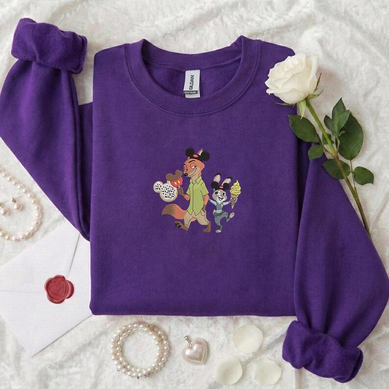 May include: A purple sweatshirt with a crew neck and long sleeves. The sweatshirt features an embroidered design of two cartoon characters, a fox and a rabbit, holding ice cream cones. A white rose and a pearl necklace are in the background.