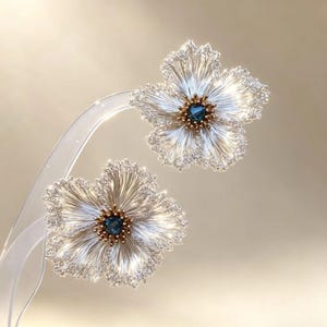 May include: A pair of flower-shaped earrings featuring silver-toned wire petals with intricate, textured edges. Each earring has a central cluster of small gold-toned beads surrounding a single, faceted, deep blue gemstone at the very center.