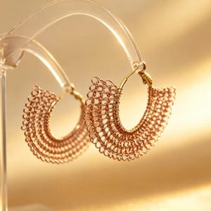Peut inclure: Une paire de boucles d'oreilles créoles dorées présentant un design en maille délicat en demi-cercle, composé de fines boucles de fil entrelacées. Les boucles d'oreilles ont une forme incurvée en éventail rayonnant depuis un fil central.
