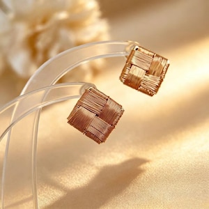 May include: A pair of square-shaped stud earrings crafted from thin, metallic copper-colored wire woven into a tight, basket-weave pattern. The earrings are displayed on a clear, curved plastic stand, showcasing the intricate texture of the metal wire.