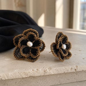 May include: A pair of stud earrings shaped like flowers, featuring black thread petals with metallic gold wire edges and a single white pearl at the center of each bloom. The intricate design highlights the contrast between the dark thread and gold.