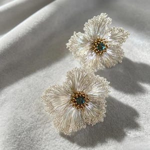 May include: A pair of flower-shaped earrings crafted from fine silver-toned wire, featuring delicate, fringed petals and a central cluster of small gold-toned beads surrounding a single, deep blue gemstone at the center of each floral piece.