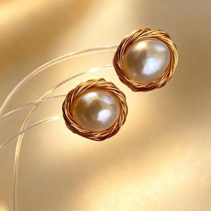 May include: A pair of stud earrings featuring round, lustrous white pearls centered within a circular frame of thin, coiled gold-toned wire. The design creates a nest-like appearance around each pearl, highlighting the smooth, reflective surface.