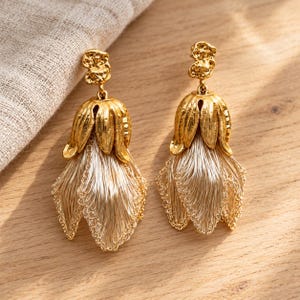 Peut inclure: Une paire de boucles d'oreilles pendantes dorées avec un clou texturé de forme organique. Sous le clou, une coupelle métallique dorée en forme de fleur retient un groupe de fins fils métalliques clairs formant un pompon à franges en pétales.