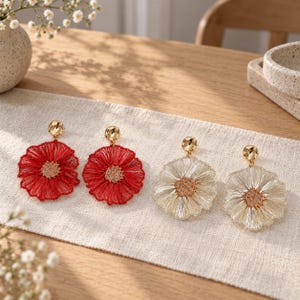 May include: Two pairs of flower-shaped dangle earrings. One pair features vibrant red thread petals with a gold-tone center, while the other pair features cream-colored thread petals with a gold-tone center. Each earring has a gold-tone stud top.