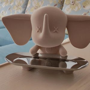 May include: A light pink elephant figurine with crossed-out eyes and a trunk, standing on a silver tray. The elephant has large ears and small arms. The tray has a reflective surface. The figurine is on a wooden table.