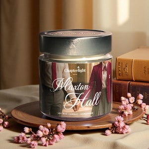 Può includere: Una candela in barattolo di vetro con coperchio argentato, su un piatto di legno. L'etichetta recita "Maxton Hall" con un'immagine di due persone che si tengono per mano. Fiori rosa e libri circondano la candela.