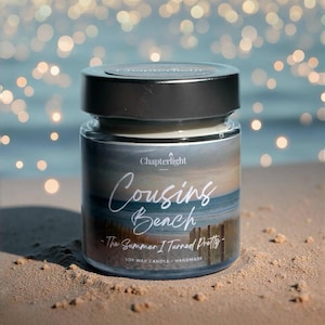 Puede incluir: Una vela en tarro de cristal con tapa plateada, etiquetada como "Cousins Beach - The Summer I Turned Pretty". La vela tiene un diseño de escena de playa y está hecha de cera de soja. El fondo es una playa arenosa con un océano borroso.