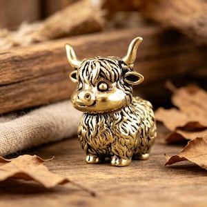 May include: A small, metallic gold-coloured figurine of a Highland cow. The sculpture features a textured, shaggy coat, two prominent curved horns, and large, round eyes. The figure is depicted in a standing position with a detailed, realistic face.