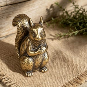 Può includere: Una dettagliata statuina di scoiattolo in ottone con finitura strutturata. Lo scoiattolo è in posizione seduta, tenendo le zampe unite. La statuina ha una coda folta e lineamenti dettagliati. Lo sfondo comprende una superficie di iuta e vegetazione.