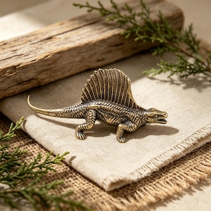 May include: A detailed brass Dimetrodon figurine with a large dorsal fin, textured skin, and open mouth. The dinosaur is posed on a tan linen cloth, with a wooden plank and greenery in the background. A decorative item.