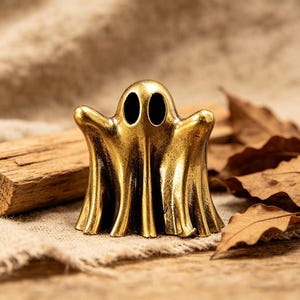 May include: A small, metallic gold-colored ghost figurine with a smooth, polished finish. The ghost features two prominent, hollow, oval-shaped black eye sockets and a draped, flowing body shape that flares out at the base, standing upright.
