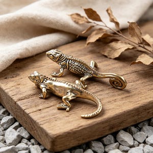 Könnte beinhalten: Zwei Messing-Echsenfiguren mit detaillierten Schuppen und eingerollten Schwänzen liegen auf einer Holzoberfläche. Die Eidechsen sind in verschiedenen Winkeln positioniert, was ihr kompliziertes Design hervorhebt. Der Hintergrund umfasst getrocknete Blätter und ein neutral gefärbtes Tuch.