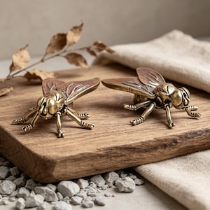 Può includere: Due figurine di mosche in ottone con ali e zampe dettagliate sono esposte su un tagliere di legno. Le mosche hanno un'estetica vintage e sono posizionate vicino a foglie secche e un panno di colore neutro.