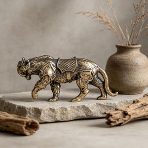 Könnte beinhalten: Eine detaillierte Messingfigur eines Tigers, verziert mit Rüstung und Sattel. Die Skulptur ist in einer Laufhaltung dargestellt und zeigt komplizierte Details. Der Tiger steht auf einer Steinoberfläche, mit einem neutralen Hintergrund und einer Vase mit getrockneten Pflanzen.