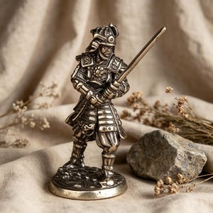 May include: A detailed silver figurine of a samurai warrior holding a katana. The samurai is in ornate armour and helmet, on a circular base. The background has neutral fabric and a small rock. The figurine is approximately 10 cm tall.