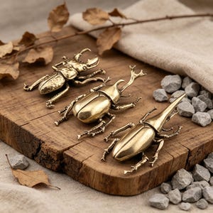 May include: Three brass beetle figurines are arranged on a rustic wooden board. The beetles vary in size and design, with intricate details. The background includes dried leaves, a beige cloth, and small gray stones, creating a natural aesthetic.