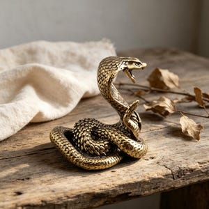 May include: A brass cobra figurine in a coiled, upright position with its hood flared and mouth open. The metal surface features detailed, textured scale patterns across the entire body, reflecting light with a metallic gold-toned finish.
