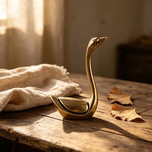May include: A polished brass figurine shaped like a swan with a long, slender neck and a smooth, curved body. The metallic surface reflects light, highlighting the detailed contours of the wings and the pointed beak of the bird sculpture.