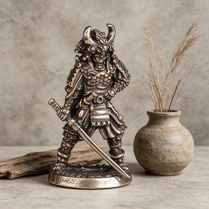 May include: A detailed bronze-colored samurai warrior figurine, holding a katana sword. The figure is clad in ornate armor and a horned helmet. A small, textured vase with dried grass sits to the right. The overall aesthetic is detailed and artistic.