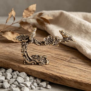 May include: A bronze-colored decorative sculpture of two birds perched on a flowering branch. The sculpture is set on a wooden surface with small gray stones at the base. The birds and flowers are intricately detailed.
