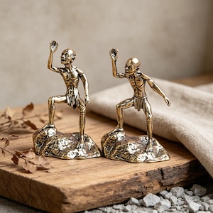 May include: Two bronze-coloured figurines of a creature with a bald head, standing on a rock base. The figures are posed with one arm raised. The figurines are displayed on a wooden surface with a neutral background.