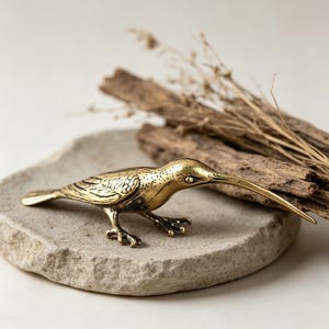 May include: A detailed brass bird figurine with a long, slender beak and intricate feather detailing. The bird is posed on a textured, light gray stone, with dried twigs and wood pieces in the background. The figurine is approximately 6 inches long.