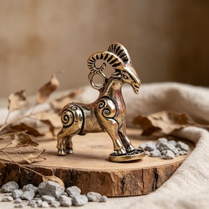May include: A bronze-colored ram figurine with detailed horns and decorative swirls on its body. The ram stands on a small, round base, placed on a wooden slice with small rocks around it. The background is a neutral tone.