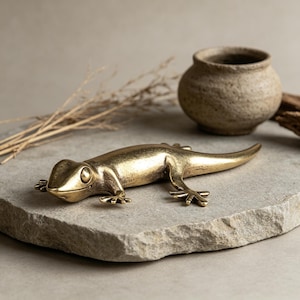 Könnte beinhalten: Eine goldfarbene Metall-Echsenfigur mit detaillierten Beinen und einem langen Schwanz. Die Eidechse liegt auf einer grauen Steinoberfläche. Ein kleiner, strukturierter, brauner Keramiktopf befindet sich im Hintergrund.