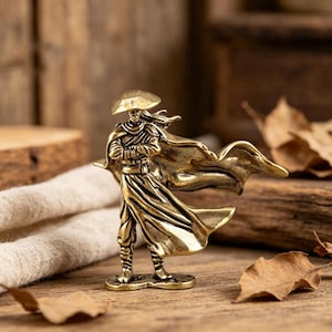 May include: A brass-colored metal figurine of a standing figure wearing a wide-brimmed conical hat and a long, flowing cloak that extends to the side. The figure has arms crossed over the chest and wears layered robes with detailed folds.