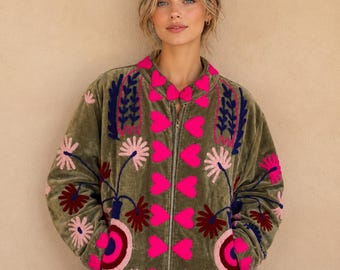 May include: An olive green velvet bomber jacket featuring a front zipper closure. The jacket is decorated with vibrant embroidery in shades of hot pink, dark blue, and light pink, depicting floral patterns and a vertical row of heart shapes.