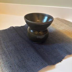 Peut inclure: Un petit bol en céramique sombre et arrondi, doté d'un large bord évasé et d'une finition lisse et brillante. Le bol présente un vernis profond et moucheté noir et vert foncé qui reflète la lumière, créant un éclat subtil sur son corps bombé.
