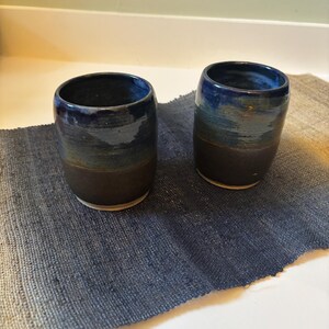 May include: Two cylindrical ceramic cups featuring a two-tone glaze. The upper portion of each cup is a glossy deep blue, while the lower portion is a matte dark brown. The cups have a smooth, rounded rim and a simple, handleless design.