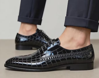 Puede incluir: Un par de zapatos de vestir negros con cordones, acabado brillante y textura de piel de cocodrilo. Los zapatos tienen un tacón bajo, punta redondeada y cordones negros finos, combinados con pantalones oscuros con dobladillo.
