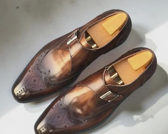 Puede incluir: Un par de zapatos de cuero marrón pulido con cierre de hebilla y diseño de ala con perforaciones decorativas en la puntera. Cada zapato incluye una hebilla de metal plateado y una horma de madera de color claro en su interior.