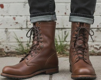 Puede incluir: Un par de botas de cuero marrón a media pantorrilla con cordones oscuros, ojales metálicos y punta redondeada. Las botas se llevan con pantalones vaqueros de color gris oscuro que tienen un dobladillo visible en la parte inferior.