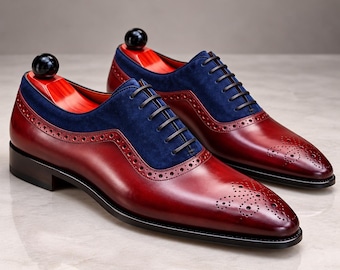 Puede incluir: Un par de zapatos oxford con una base de cuero rojo oscuro y un panel superior de ante azul marino en contraste. Estos zapatos incluyen cordones de color marrón oscuro, detalles brogue a lo largo de las costuras y una puntera decorada.