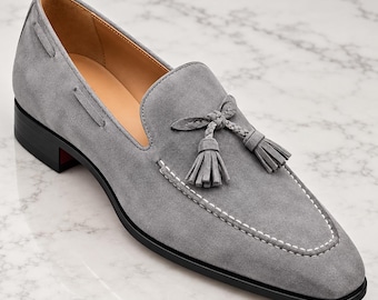 Puede incluir: Un mocasín de ante gris con un detalle de borlas trenzadas en el empeine, costuras blancas en contraste a lo largo de la puntera, un forro interior de cuero color canela y una suela negra con un tacón bajo de bloque.