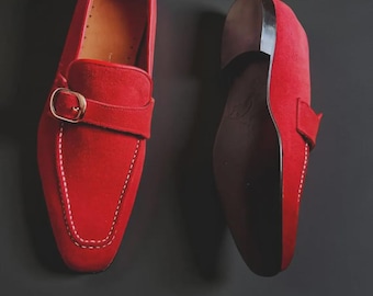 Puede incluir: Un par de mocasines de ante rojo brillante con una tira única sobre el empeine y una hebilla de metal dorado. Los zapatos tienen una puntera cuadrada, costuras blancas en contraste a lo largo del empeine y una suela negra lisa.