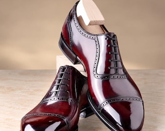 Puede incluir: Un par de zapatos oxford de cuero color burdeos pulido con diseño de puntera y detalles decorativos de brogue a lo largo de las costuras. Los zapatos tienen cordones negros, tacón apilado y una horma de madera clara en uno.