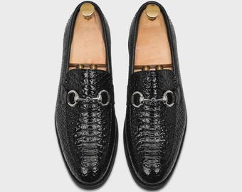 Puede incluir: Un par de mocasines negros con un acabado brillante y texturizado que recuerda a la piel de reptil. Cada zapato está adornado con un detalle de bocado de metal plateado en el empeine e incluye una horma de madera clara en su interior.