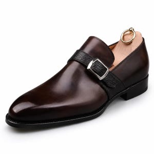 Puede incluir: Un zapato de cuero marrón oscuro con una sola hebilla, acabado liso en la puntera y una tira de cuero negro texturizado sobre el empeine. El zapato cuenta con una hebilla de metal plateado y una horma de madera clara con un anillo dorado.