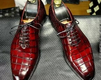 Puede incluir: Un par de zapatos oxford de color rojo oscuro con una textura brillante de piel de cocodrilo, cordones negros y punta afilada. Cada zapato está equipado con una horma de plástico negro para mantener su forma y estructura.