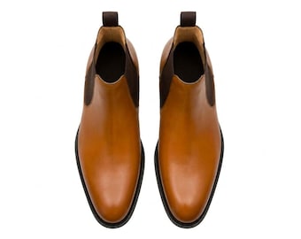 Puede incluir: Un par de botas Chelsea de cuero color canela con acabado liso, punteras redondeadas y paneles laterales elásticos de color marrón oscuro. Cada bota incluye una trabilla marrón oscuro en el talón y una suela negra, vista desde arriba.