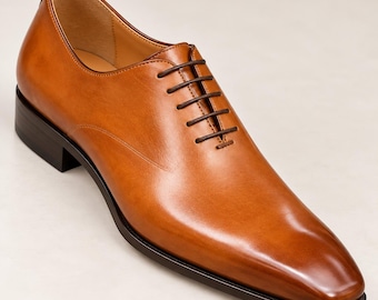 Puede incluir: Un zapato oxford de cuero color canela con acabado liso, punta redondeada y cordones marrón oscuro. El zapato tiene un tacón bajo y una suela marrón oscuro, con un forro interior beige visible en la abertura del cuello del zapato.