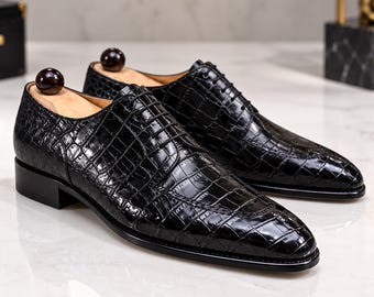 Puede incluir: Un par de zapatos oxford negros con textura de piel de cocodrilo brillante, cordones negros finos, punta afilada y tacón bajo. Cada zapato contiene una horma de madera con un pomo esférico de color marrón oscuro en el talón.