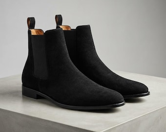 Puede incluir: Un par de botas Chelsea de ante negro con paneles laterales elásticos, punta redondeada y una trabilla en el talón. Las botas tienen un tacón bajo y una suela oscura, presentando una silueta limpia y sencilla que llega al tobillo.