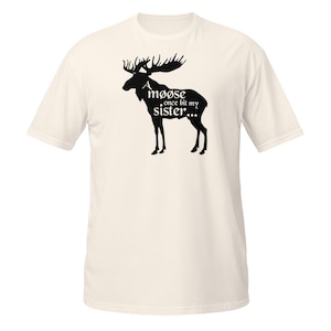 Può includere: Una t-shirt a maniche corte color crema con la sagoma nera di un alce. Il testo "A møøse once bit my sister..." è stampato in nero sul corpo dell'alce, utilizzando caratteri stilizzati con barre diagonali sulla lettera o.